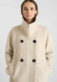 Chaqueta de forro polar beige con cuello alto, botones negros de doble pecho y bolsillos laterales. Textura suave, corte relajado, cierre de cremallera.