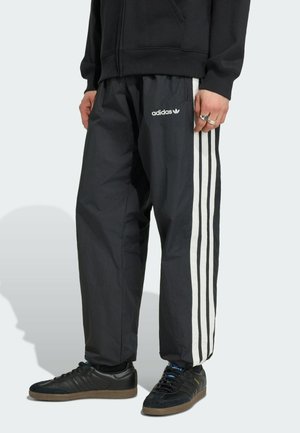 Osoba nosící černé tepláky Adidas s bílými pruhy po stranách a černé tenisky Adidas s gumovou podrážkou.