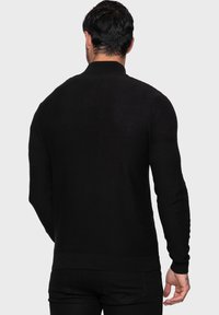 Mann, der von hinten zu sehen ist, trägt einen schwarzen Pullover mit langen Ärmeln und strukturierten Oberärmeln sowie schwarze Hosen vor einem einfarbigen Hintergrund.