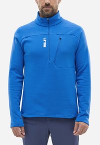 Mann in einem leuchtend blauen Fleece-Pullover mit einem Viertel-Reißverschluss und einer Reißverschlusstasche auf der Brust, kombiniert mit einer dunklen Hose.