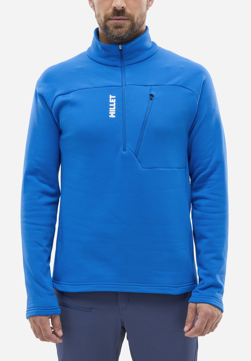 Mann in einem leuchtend blauen Fleece-Pullover mit einem Viertel-Reißverschluss und einer Reißverschlusstasche auf der Brust, kombiniert mit einer dunklen Hose.