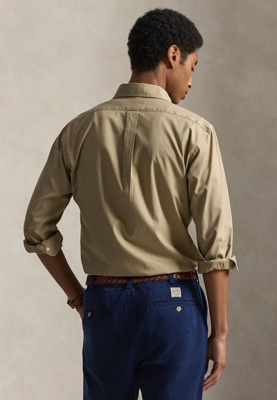 Άνδρας με σηκωμένα μανίκια φορώντας μπεζ πουκάμισο με κουμπιά, μπουρνιασμένο μέσα σε σκούρο μπλε παντελόνι με πλεκτή καφέ ζώνη, στέκεται μπροστά σε ουδέτερο φόντο.