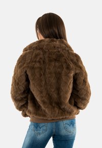 Giacca in ecopelliccia marrone con design trapuntato, colletto alto e vita elastica. Abbinata a jeans in denim blu con cuciture decorative.