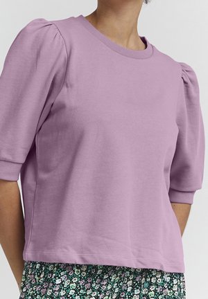 Kobieta ubrana w lawendową bluzkę z bufkami, zestawioną z spódnicą w kwiatowy wzór z fioletowymi, zielonymi i białymi kwiatami.