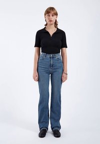 Polo negro de canalé, vaqueros azules acampanados de talle alto, zapatos negros. La modelo está de pie, erguida, contra un fondo liso. Atuendo sencillo y casual.