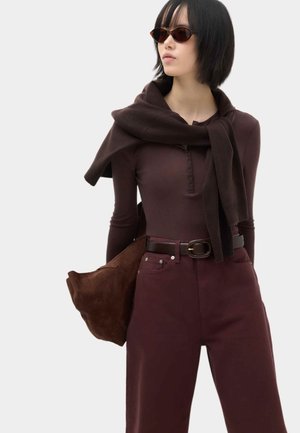 Donna con capelli corti e neri che indossa occhiali da sole marrone scuro, maglia a maniche lunghe, pantaloni, un maglione adagiato sulle spalle e porta una borsa marrone.