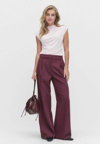 Vaaleanpunainen lyhythihainen toppi, jossa on tiukka siluetti, yhdistetty tumman burgundin värisiin leveälahkeisiin housuihin. Malli pitää käsissään ruskeaa narupussukkaa.