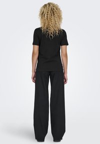 Mujer con largo cabello rizado rubio, vistiendo una camiseta negra de manga corta y pantalones anchos negros, de pie de espaldas sobre un fondo liso.