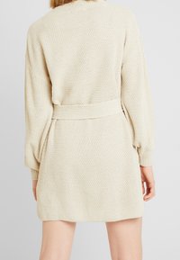 Stickad beige cardigan med avslappnad passform, texturerat tyg, långa ärmar och ett matchande bälte i midjan. Enkelt, mångsidigt design.