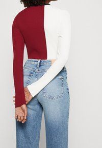 Top corto a coste con maniche lunghe; metà bordeaux, metà bianco. Abbinato a jeans chiari a vita alta. La vista posteriore mostra le mani incrociate.