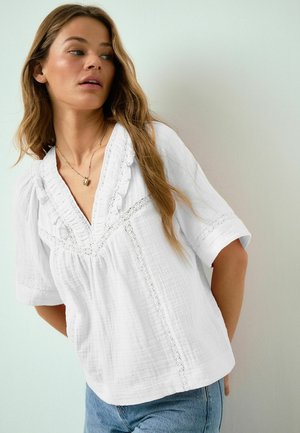 Young woman with long wavy hair wearing a white textured blouse with lace details and blue jeans, looking to the side against a plain background.