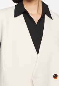 Gilet blanc en tissu texturé, avec un décolleté en V, un devant double boutonnage et un grand bouton en écaille de tortue. Chemise noire en dessous.