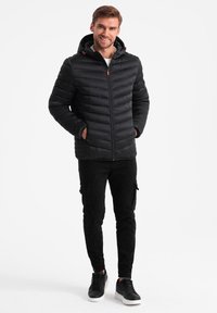 Schwarze Pufferjacke mit Kapuze, vertikales, gestepptes Design, Frontreißverschluss und roten Akzenten. Kombiniert mit schwarzen Cargo-Hosen und Turnschuhen.