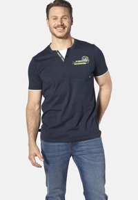 Marineblaue Kurzarm-Polohemd mit Henley-Ausschnitt, kontrastierenden weißen Akzenten und einem gedruckten Design auf der Brust. Getragen mit blauen Jeans.