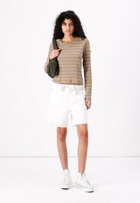 Langermet brun og beige stripete topp paret med hvite, slitte denimshorts og hvite joggesko, holder en oliven grønn veske.