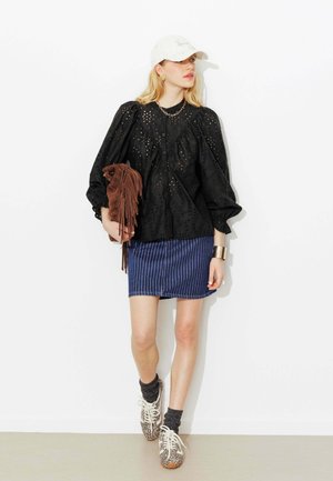 Femme portant un chemisier noir à œillets, une jupe rayée bleue, une casquette blanche, des baskets imprimé léopard, tenant un sac marron à franges, debout contre un mur blanc.