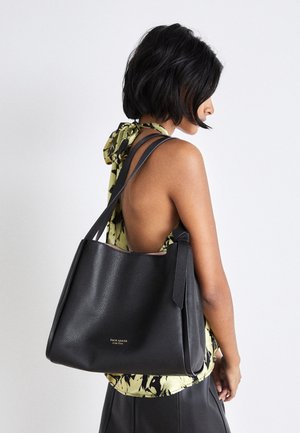 Femme aux cheveux noirs courts portant un haut vert à motifs et dos nu, avec un grand sac cabas en cuir noir Kate Spade sur l'épaule.
