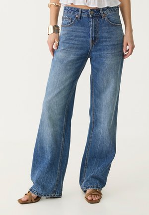 Frau trägt blaue High-Waist-Weitbein-Jeans, weißes Crop-Top, goldene Armbänder, Ringe und braune Sandalen mit offenen Zehen, steht vor weißem Hintergrund.