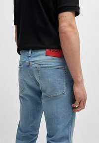 Ljusblå denimjeans med en röd läderetikett med "HUGO" på midjebandet. Designen inkluderar bakfickor och en rak passform.