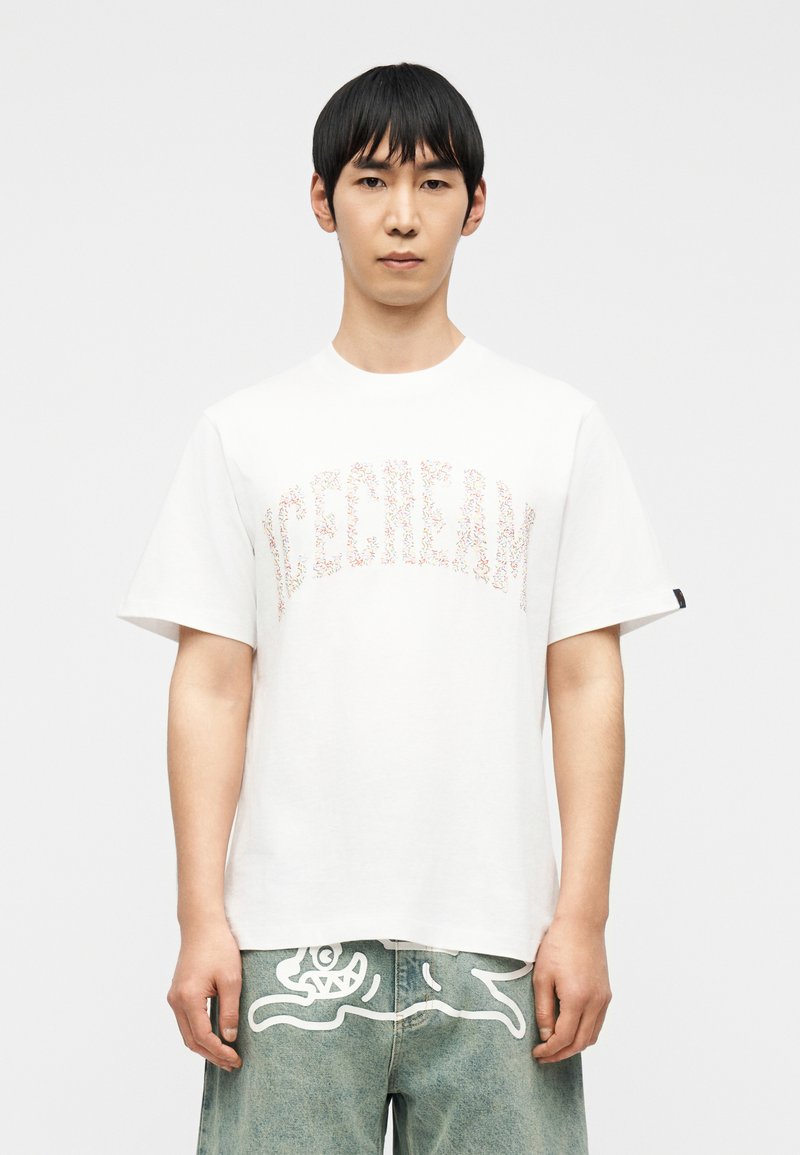 Νέος άνδρας που φοράει λευκό T-shirt με πολύχρωμο κείμενο «ICECREAM» και ανοιχτό μπλε τζιν με λευκό σχέδιο αφηρημένου πλάσματος.