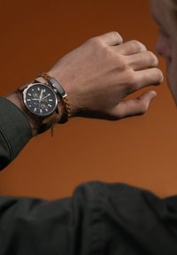 Brūna ādas pulksteņa ar melnu ciparnīcu, sudraba akcentiem un diviem apakšciparniekiem uz rokas, blakus pinumam no ādas.