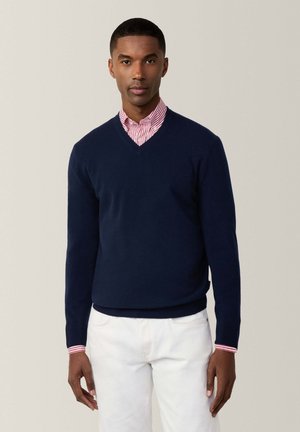Navyblauer V-Ausschnitt-Pullover aus weichem Stoff, mit langen Ärmeln und geripptem Saum. Über einem rot-weiß gestreiften Hemd mit Kragen getragen.