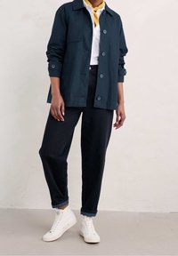 Osoba w granatowej kurtce zapinanej na guziki, białej koszuli z żółtym szalikiem, ciemnych dżinsach i białych trampkach na wysokiej podeszwie stoi na szarym podłożu.