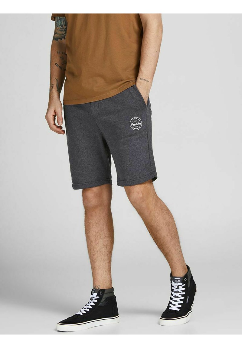 Šedé bavlněné volnočasové šortky se bočními kapsami, na levém stehně s bílým kruhovým logem. Nosené s černými vysokými teniskami.