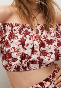 Crop top décolleté en blanc avec un motif floral dans des nuances de rouge et de marron. Tissu texturé avec un élastique froncé à la taille.