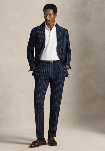 Bărbat purtând un costum bleumarin cu dungi fine, tricou polo alb, curea maro, pantofi maro cu ciucuri și ochelari, stând cu mâinile în buzunare.