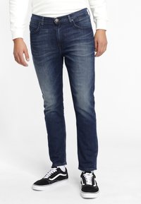 Tummansiniset farkut, joissa on kapea malli, viisi taskua ja vaalea pesu, yhdistettynä mustiin ja valkoisiin tennareihin.