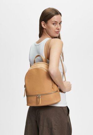 Kobieta ubrana w biały top na ramiączkach i brązowe spodnie niesie beżowy skórzany plecak na ramieniu, patrząc przez prawe ramię za siebie.