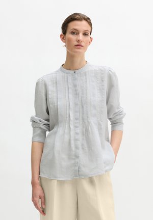 Vrouw draagt een lichtgrijze blouse met lange mouwen en verticale plooien, gecombineerd met beige wijde broekspijpen, staand tegen een effen witte achtergrond.
