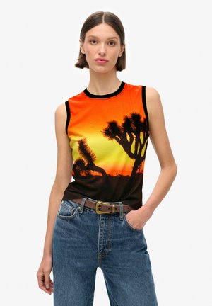 Débardeur graphique sans manches avec un design de coucher de soleil orange et jaune, comportant des contours noirs d'arbres de Joshua. Associé avec un jean en denim bleu.