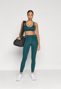 Teal sportmelltartó és magas derekú leggingszettek, texturált anyagból készült. A modell egy fekete tornazsákot tart, fehér sportcipőt visel.