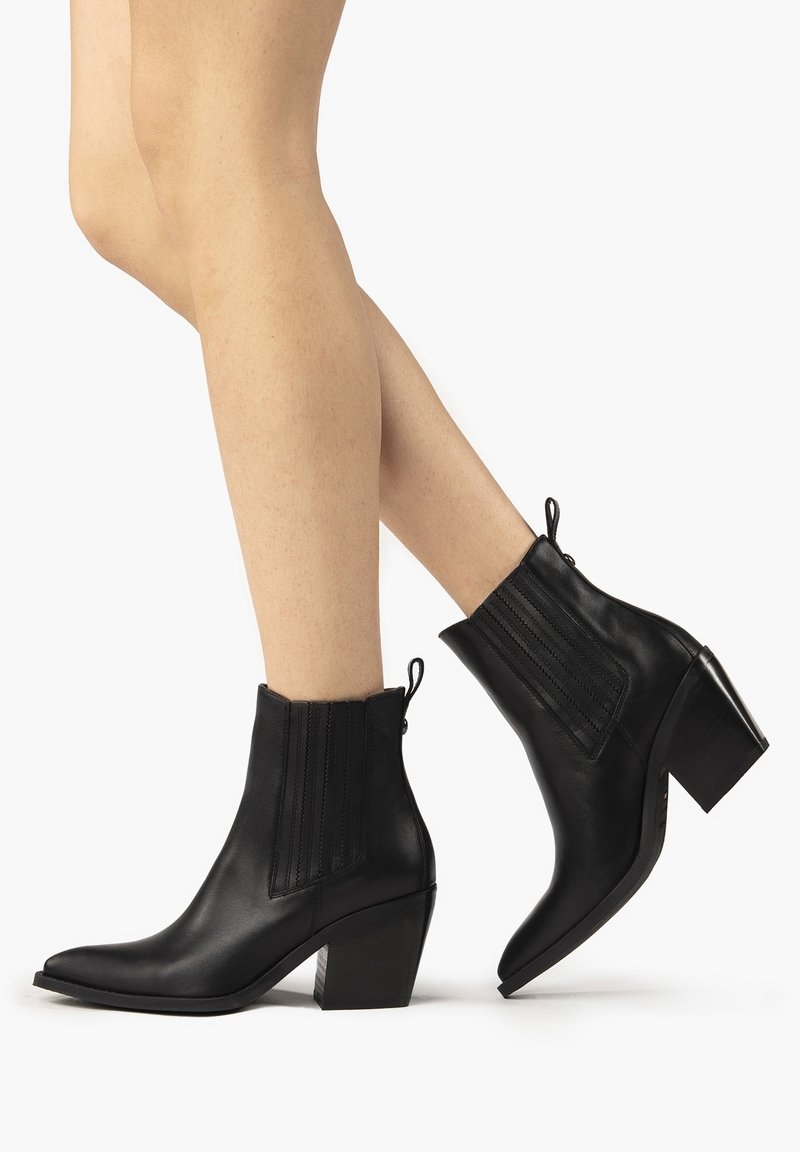 Jambes portant des bottines en cuir noir à bout pointu et talon bloc, debout sur un fond blanc uni.