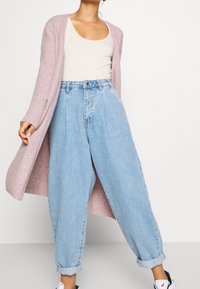 Kvinna som bär en ljusrosa lång kofta, vit linne, högmidjade, lösa ljusblå jeans och vita sneakers med blå detaljer.