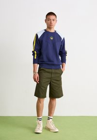 Námornícky sveter s svetlomodrými detailmi a žltými bočnými pruhmi, kombinovaný s olivovozelenými šortkami a béžovými vysokými teniskami.