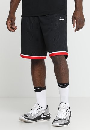 Mand iført sorte basketballshorts med røde kanter, hvide Nike-sokker og hvide og sorte Nike-sneakers, stående på en ensfarvet baggrund.