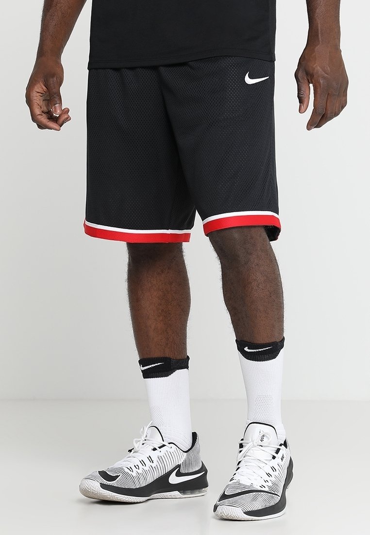 Homme portant un short de basketball noir avec des bordures rouges, des chaussettes blanches Nike, et des baskets blanches et noires Nike, debout sur un fond uni.