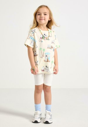 Ung pige står smilende iført en cremefarvet T-shirt med tegneseriefigurer, hvide shorts, blå sokker, hvide sneakers og farverige perlesmykker.