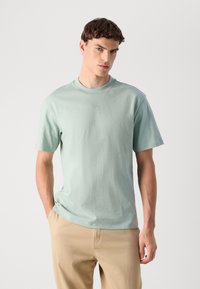 Jovem vestido com uma t-shirt de mangas curtas verde-clara e calças bege, com uma mão no bolso, de pé contra um fundo branco liso.