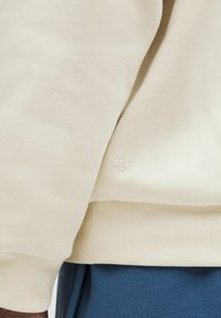 Felpa beige in tessuto texturizzato con polsini a costine, dotata di un piccolo logo sul lato inferiore. Indossata sopra pantaloni color teal.