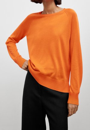 Femme portant un pull à manches longues orange vif et un pantalon noir, debout avec une main sur la hanche sur un fond uni.