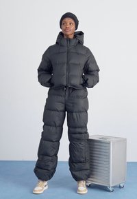 Svart puffjacka och byxor, båda med kviltad textur. Har en huva, dragsko i midjan och tan färgade sneakers bredvid en silverfärgad förvaringslåda.