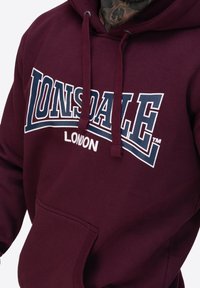 Bordowa bluza z kapturem z dużym białym i niebieskim logo Lonsdale London, sznurkami do ściągania i kieszenią z przodu. Miękka tkanina z domieszką bawełny.