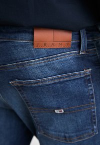 Tamno plavi traperice s smešnim kožnim oznakom "JEANS", s narančastim šavovima i dva stražnja džepa. Glatka tekstura.