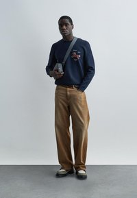 Hombre con suéter azul marino con bordado de caballo, jeans marrones de corte holgado, zapatillas blancas y verdes, y bolso bandolera sobre fondo gris liso.