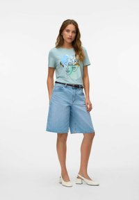 Lichtblauw t-shirt met abstracte multikleurige gezichten, gecombineerd met lichtdenim shorts en witte blokhak schoenen, met een bruine riem.