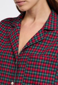 Camisa de cuadros roja y verde, con cuello con muesca, que presenta un bolsillo en el pecho y detalles en costura blanca. Textura de tela suave.